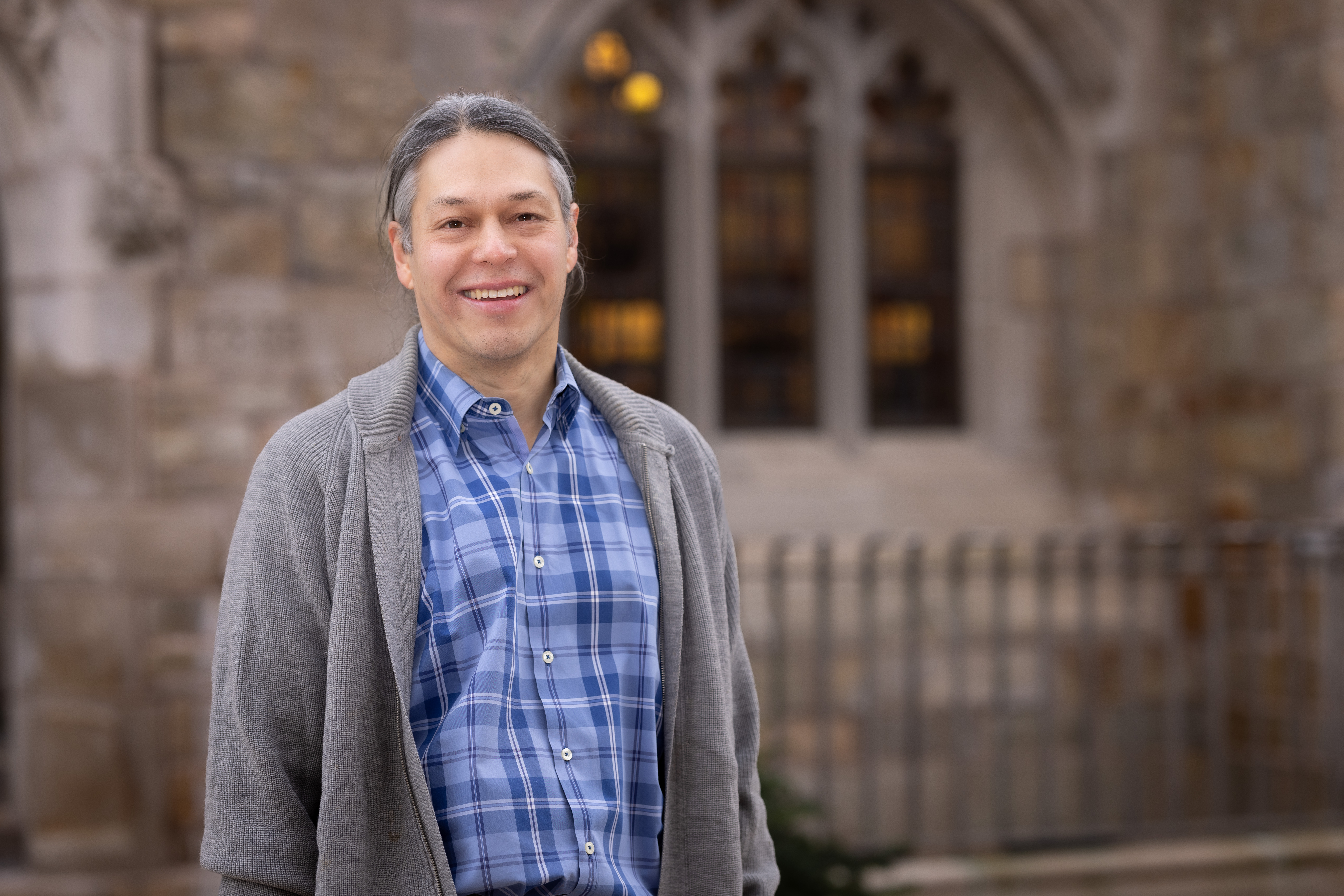 a man is smiling at the camera standing in front of a church window, he is wearing a blue checkered button down shirt with a grey sweater over it