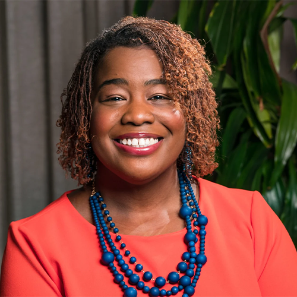 a black woman with shoulder length curly hair wearing a bright orange shirt is and blue necklace is smiling at the camera